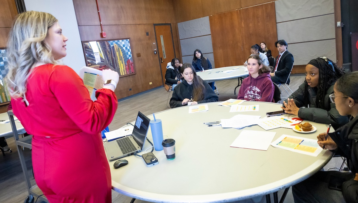 Teenage girls sitting at round tables listening to a woman giving a lecture.